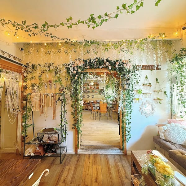A whimsical cafe interior with abundant greenery, string lights, and macrame decor, leading to a dining area.