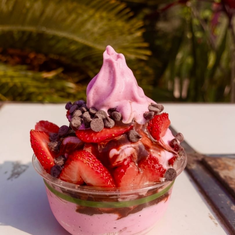 Pink soft-serve ice cream with fresh strawberries, chocolate chips, and chocolate syrup in a clear cup.