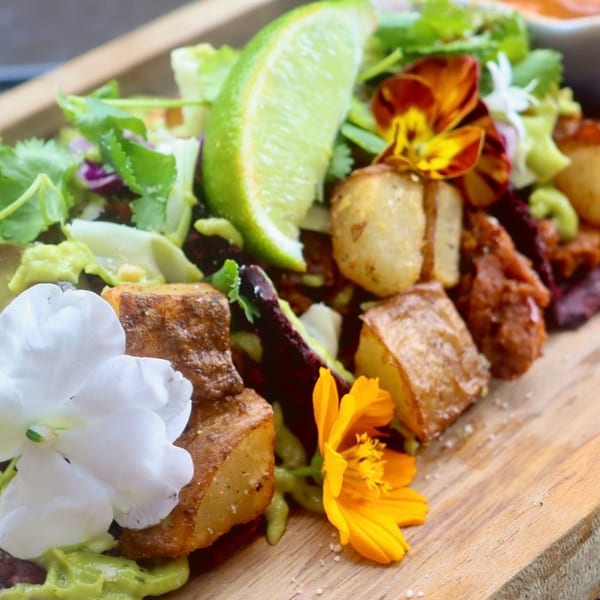 Close-up of roasted potatoes, greens, edible flowers, and a lime wedge on a wooden board.
