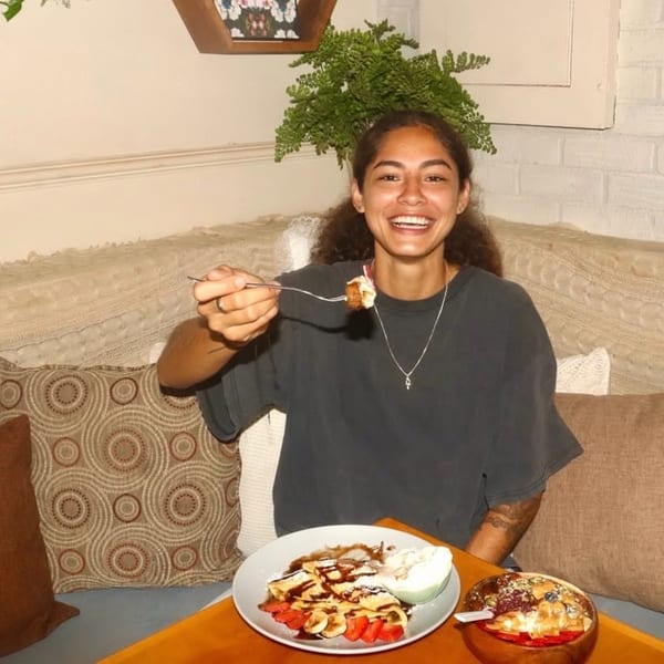A smiling woman holding a forkful of food, sitting at a table with crepes and a smoothie bowl.
