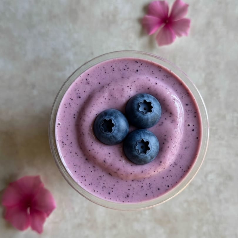 Overhead view of a purple blueberry smoothie in a glass, garnished with three fresh blueberries and pink flowers.